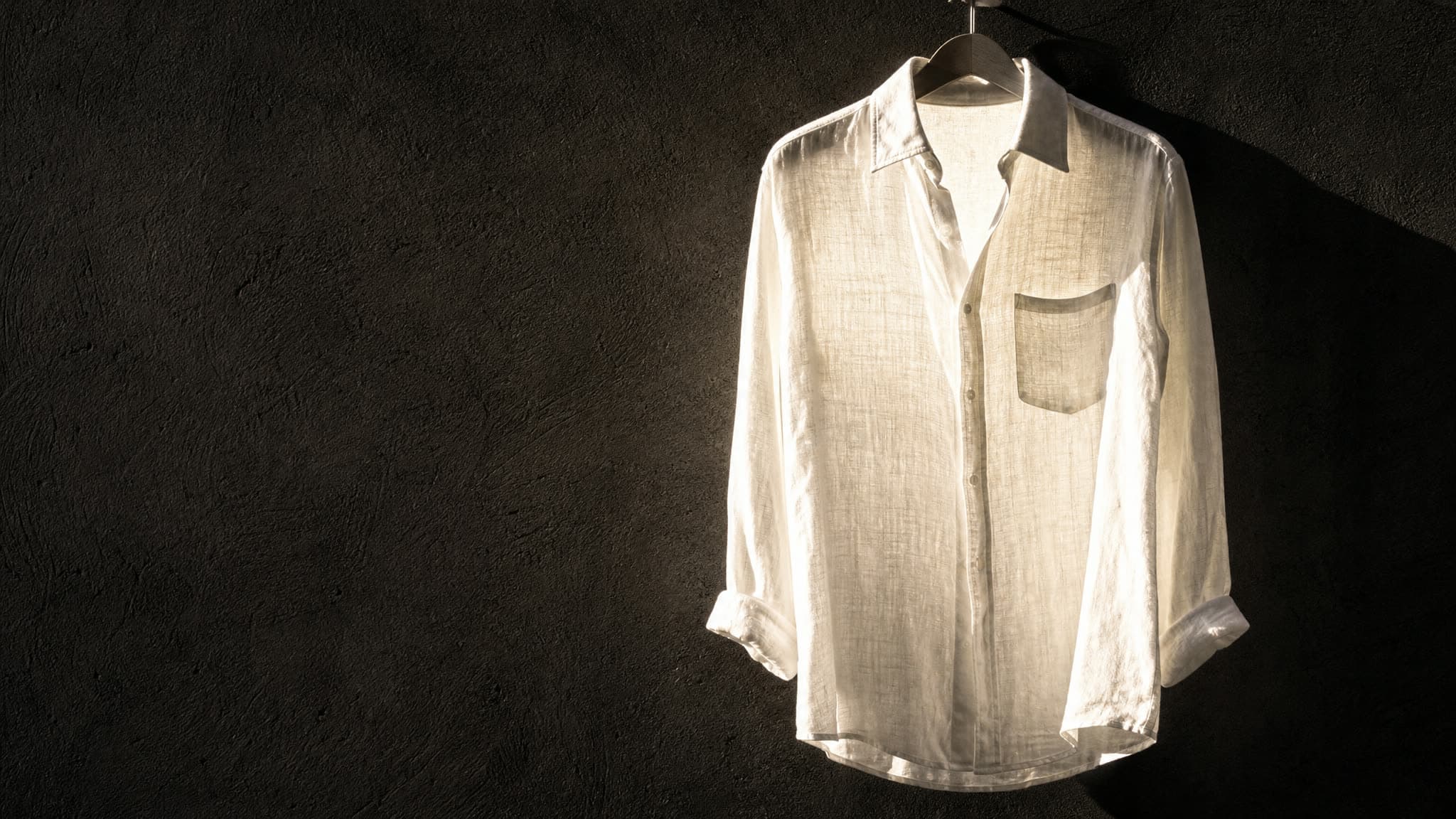 A bone-white linen shirt on a hanger against a dark wall, backlit by midday sun so the weave of the cloth is visible through it.